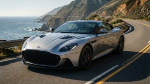 Обои Серебристый Aston Martin DBS петляет по прибрежной дороге на скалистом утесе, яркий солнечный свет.